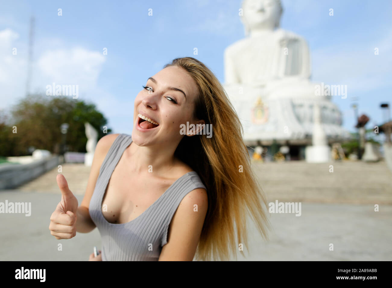 Joven Mujer rubia mostrando Thumbs up, blanca estatua de Buda en Phuket en segundo plano. Foto de stock