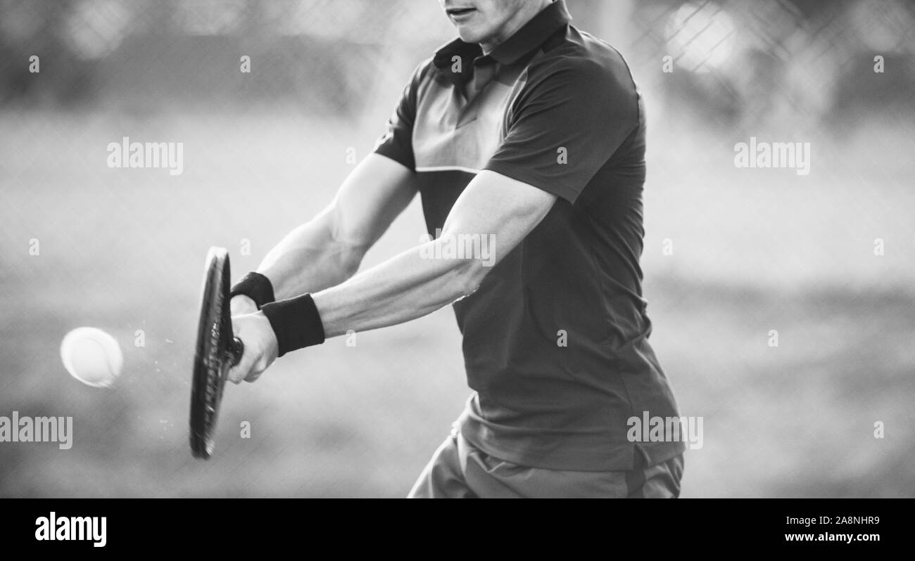 Rodada en blanco y negro de un jugador de tenis masculino golpear un revés durante un juego. Jugador profesional, jugar en la cancha de tenis. Foto de stock