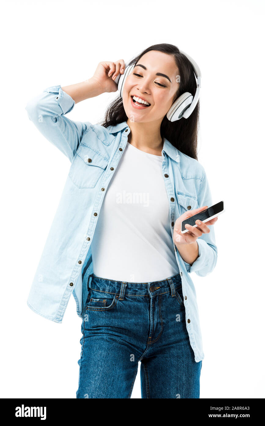 Atractiva y sonriente mujer asiática en denim shirt escuchando música y mantiene aislado en blanco del smartphone Foto de stock