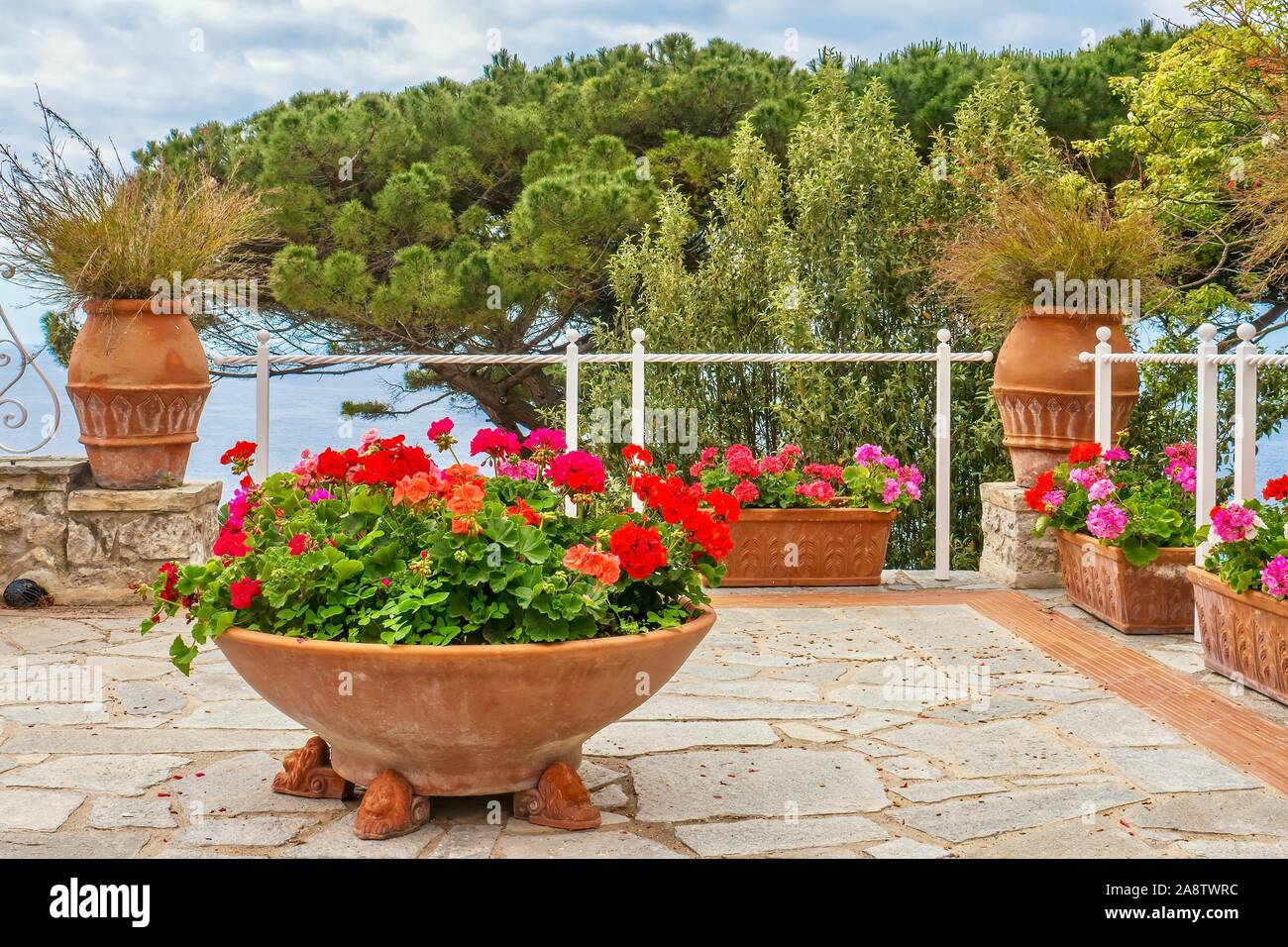 Un jardín de verano en el mar. Coloridos geranios en macetas de terracota en una terraza de piedra con vistas al mar Mediterráneo en Italia. Foto de stock