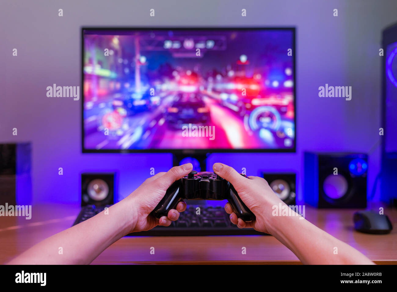El Joypad en manos. Concepto de juegos de ordenador. Ordenador con pantalla de juego en segundo plano. Luz RGB detrás del mostrador. Foto de stock