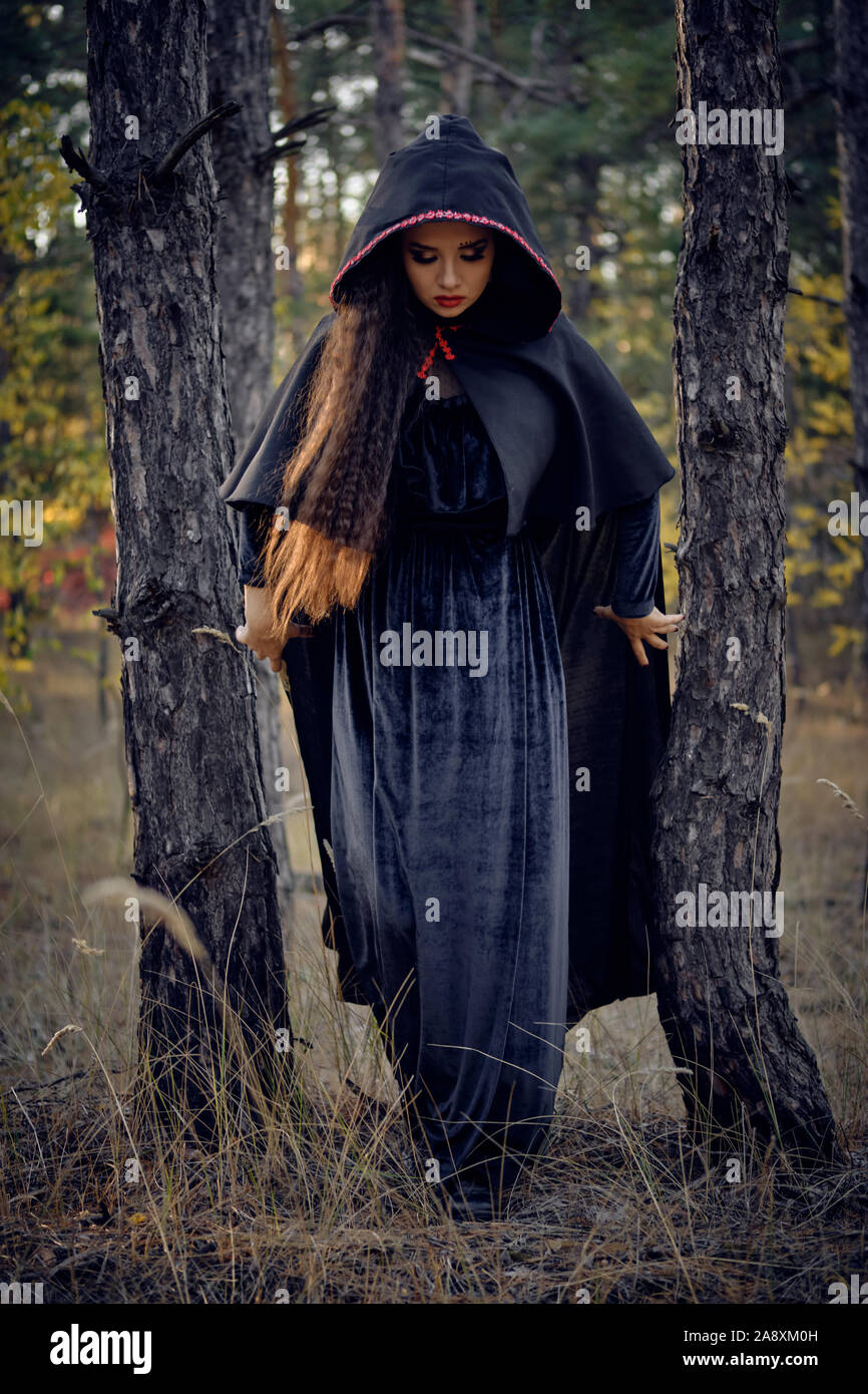 Bonita, De pelo largo, la malvada bruja en negro, vestido largo con capa y  capucha. Ella está posando en un bosque de pinos de pie entre los árboles.  Hechizos, magia un Fotografía