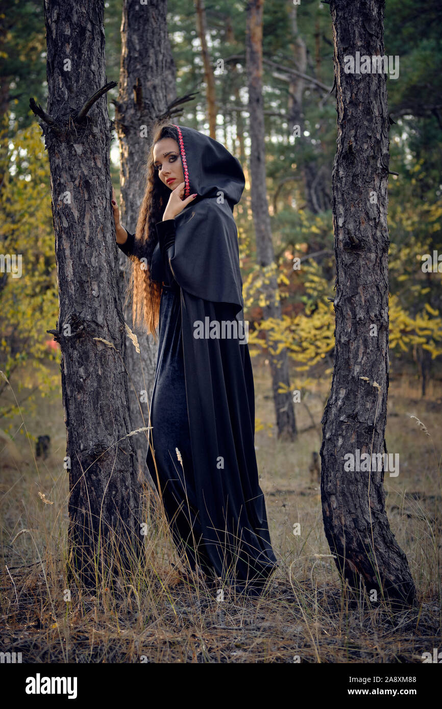 Encantador, la malvada hechicera, De pelo largo en negro, vestido largo con  capa y capucha. Ella está posando en un bosque de pinos de pie entre los  árboles. Los hechizos, ma Fotografía