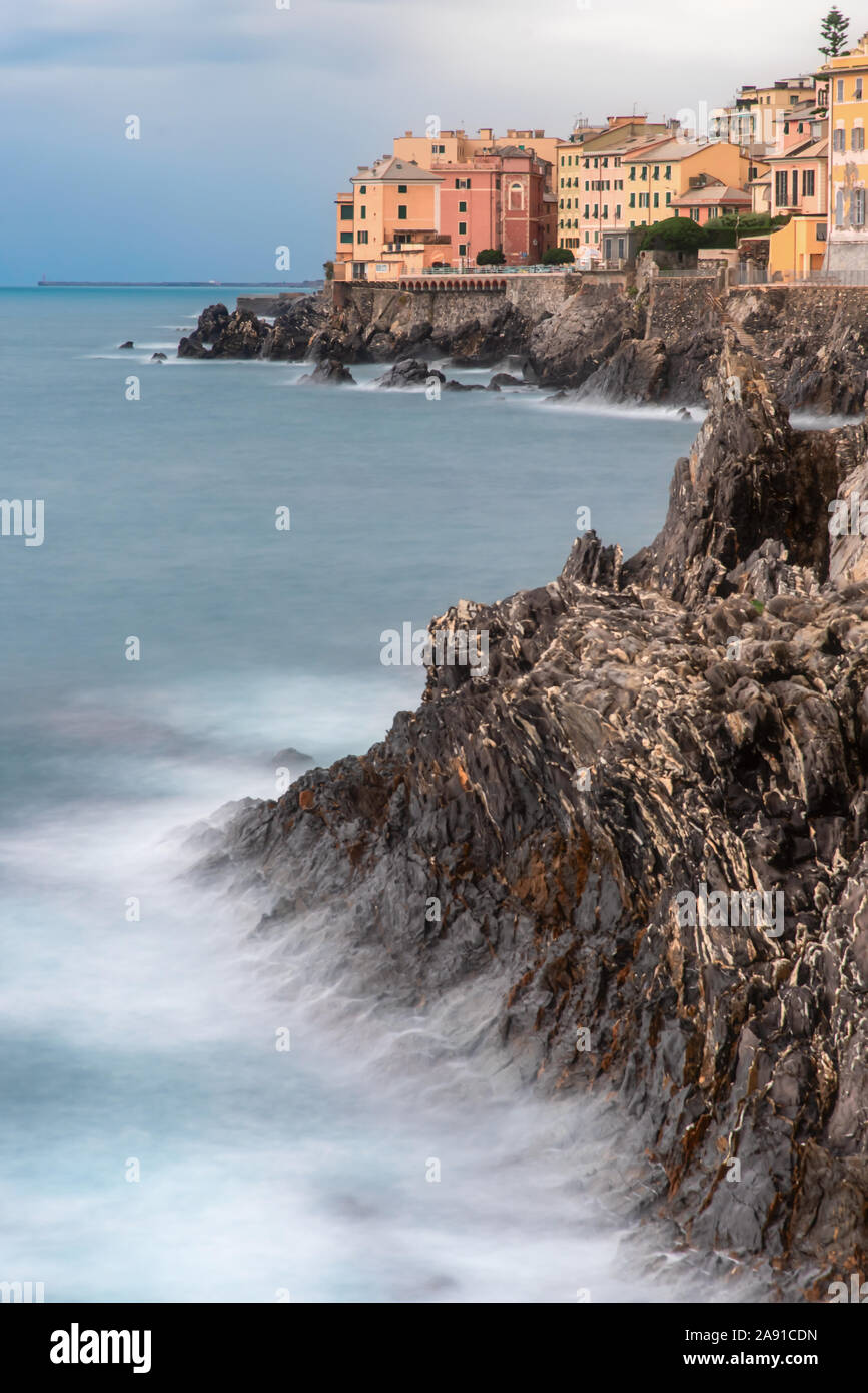 Exposición HDR de la rocosa costa y olas rompiendo en los acantilados de Nervi, Liguria, Italia con vistas a los coloridos edificios de la ciudad Foto de stock