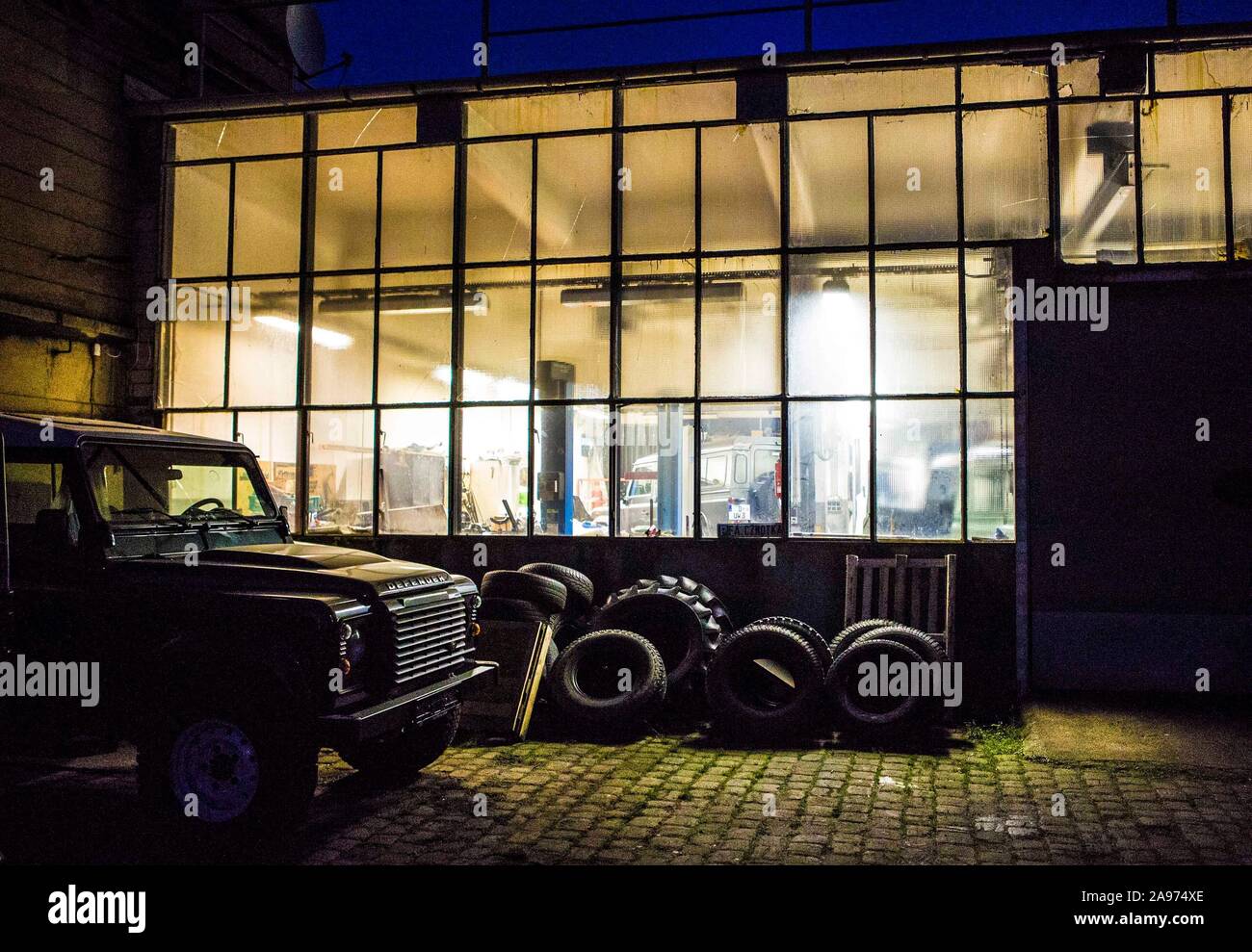 Außenansicht einer kleinen Land Rover Werkstatt im Ruhrgebiet en den Abendstunden. Den Scheiben Hinter der Halle sieht man im Licht die Autos, die dor Foto de stock