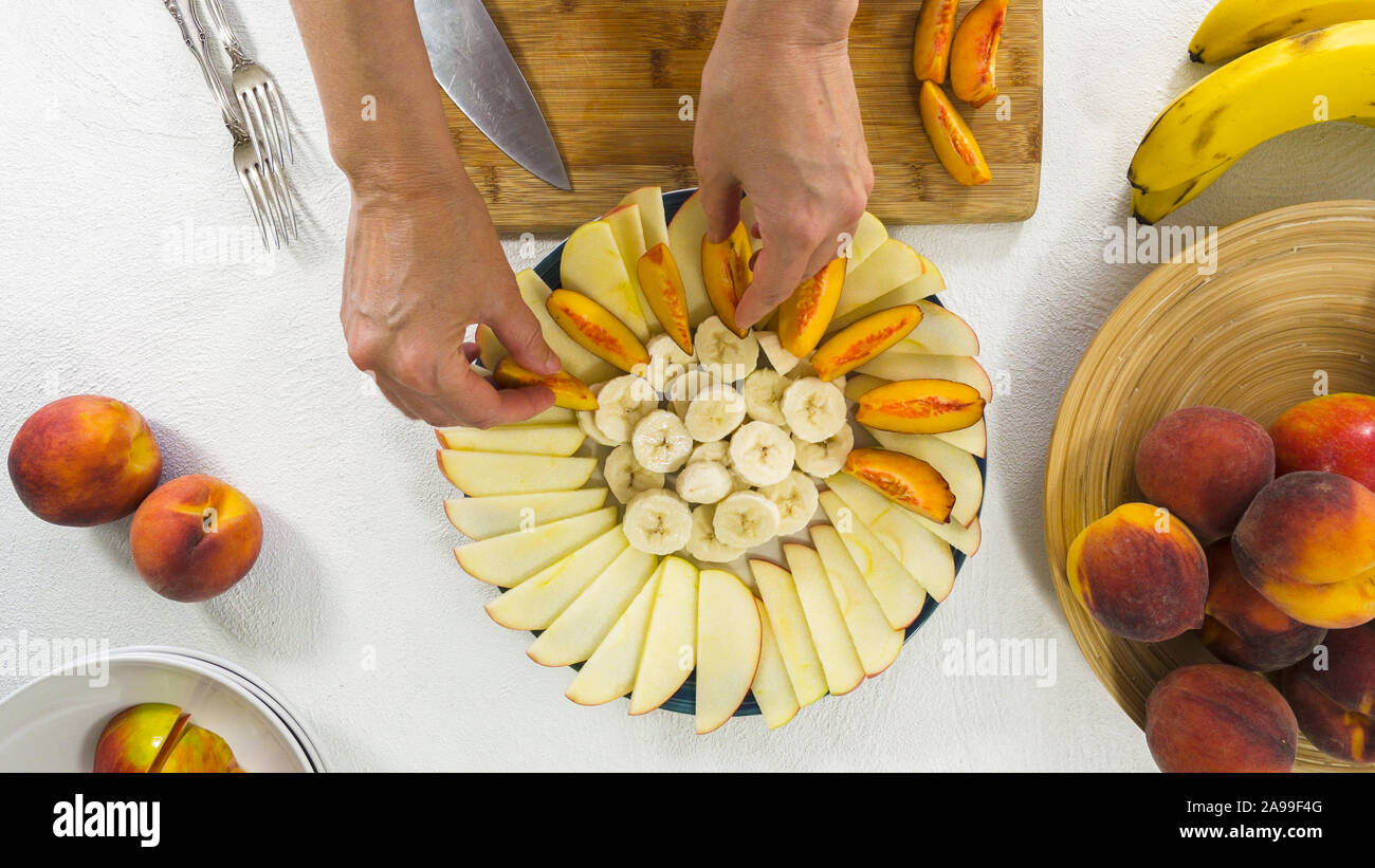 Las manzanas, plátanos y melocotones en rodajas sobre un plato. Mujer de manos, vista superior sobre fondo blanco. Ensalada de fruta, fruta, postres Foto de stock