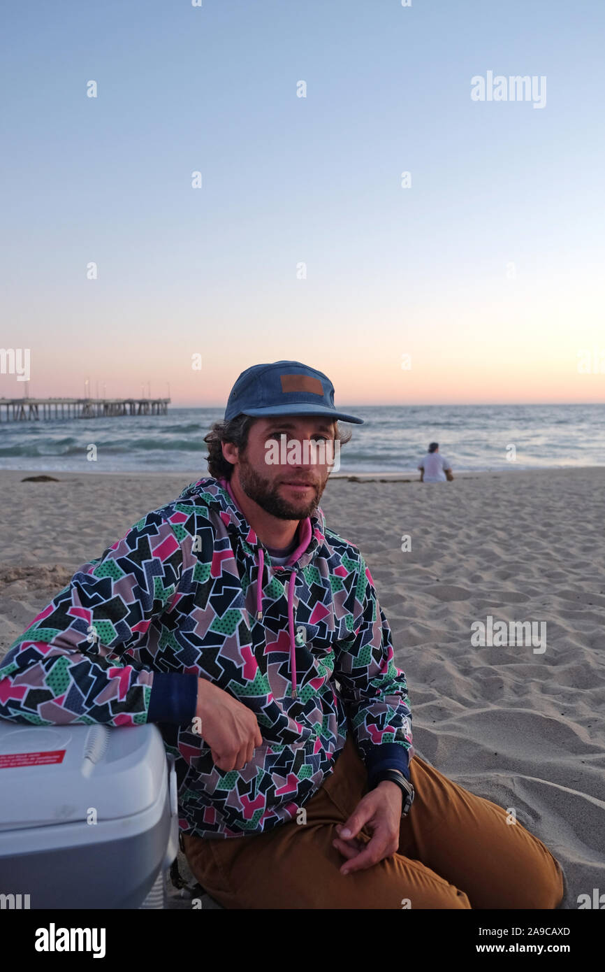El hombre llevaba una gorra con la barba y el Cabello Rizado sentado en la playa en el atardecer. Foto de stock