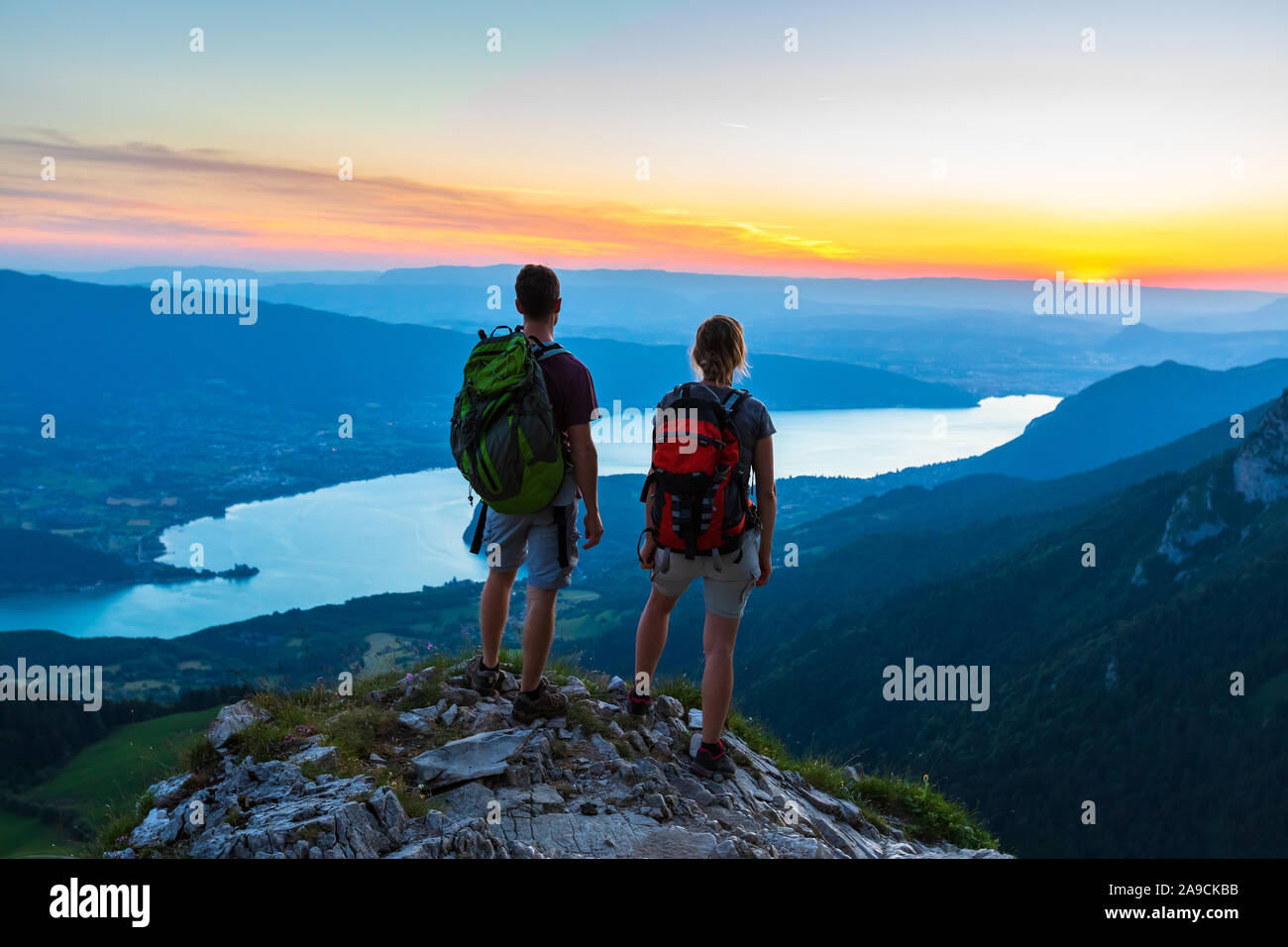 Excursionistas disfrutando de vistas panorámicas del valle con el lago al atardecer, pareja disfrutando de verano al aire libre trekking en las montañas, un estilo de vida activo, dos personas como mochilero Foto de stock