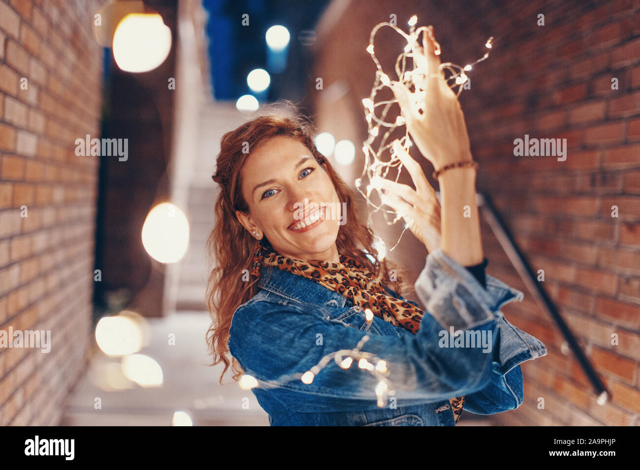 Mujer joven feliz jugando con las luces de hadas afuera en la ciudad, gran sonrisa Foto de stock