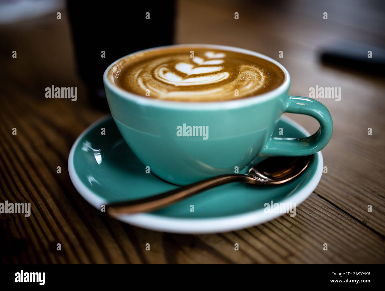 Este es un brillante taza Cappuccino con hermosas latte art en la parte superior. El aqua taza descansa sobre una mesa de madera con una cuchara de cobre acentuar la bebida. Foto de stock