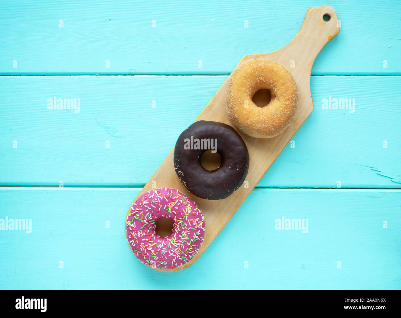 Tres diferentes donuts, chocolate donut, Rosa guinda y sin guinda sobre una tabla de cortar de madera de madera sobre un fondo azul. Foto de stock