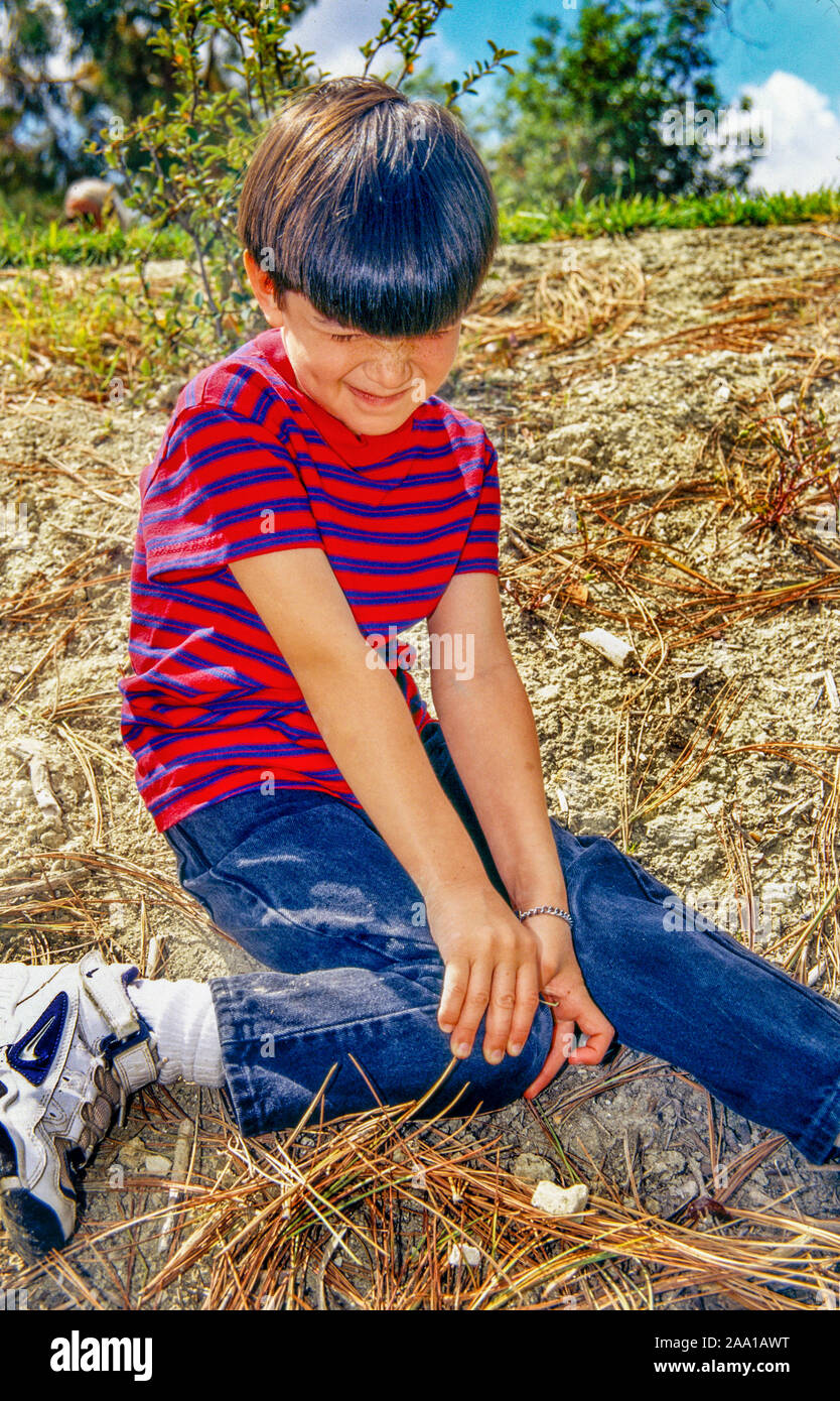 Un niño hispano winces en dolor como él roza un esguince de rodilla en Mission Viejo, California, Parque. Foto de stock