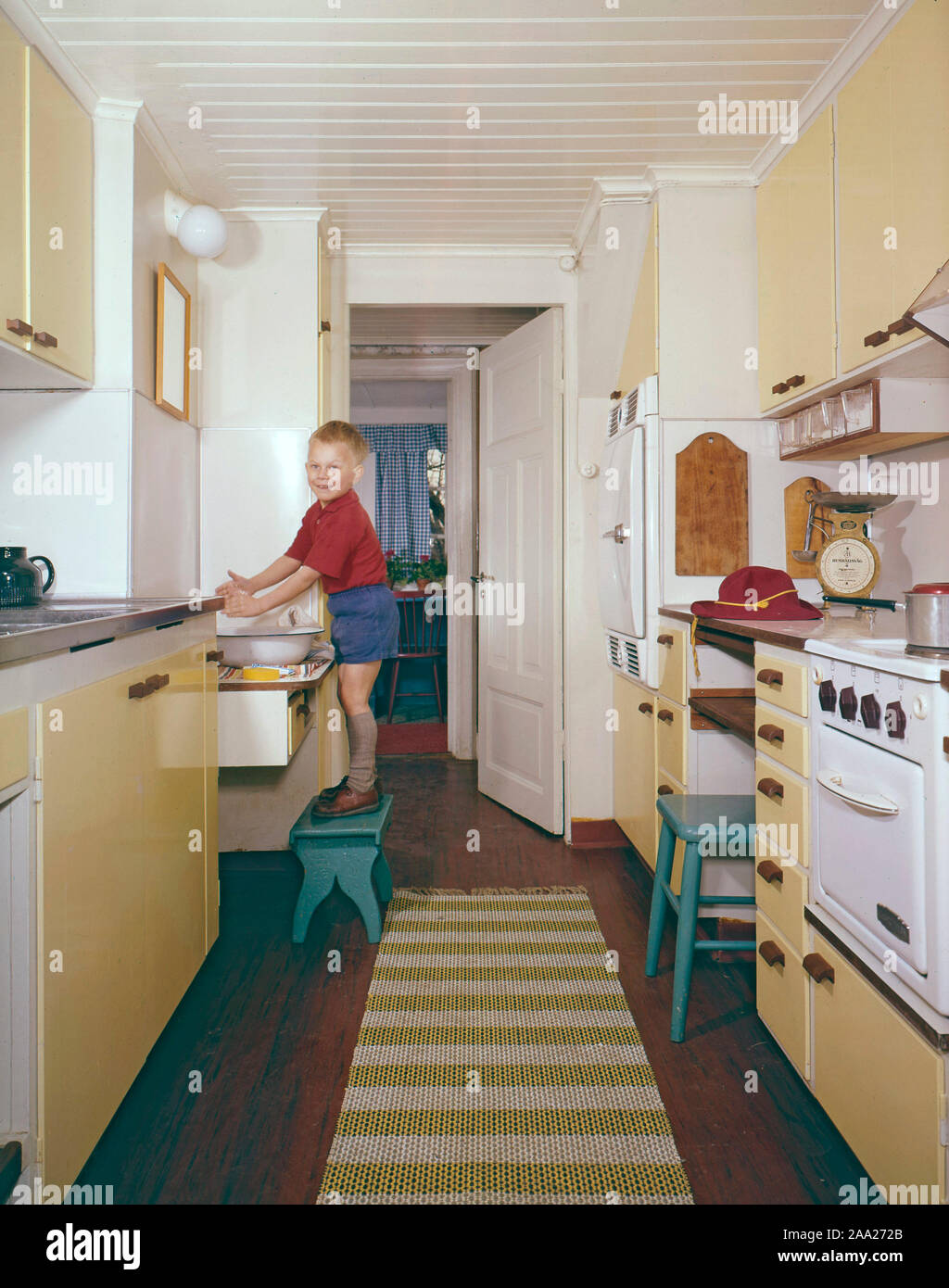 La cocina de 50S-60s. Un muchacho me s de pie sobre un taburete y se lava las manos. La cocina es típica de interior con tonos amarillos y objetos visibles del tiempo. Suecia 1950 1960 Foto de stock