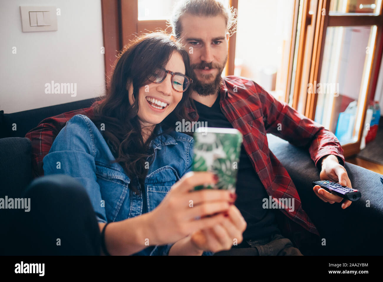 Hermosa joven feliz pareja sofá interiores domésticos teniendo selfie utilizando smartphones - tecnología, screentime, freetime concepto Foto de stock