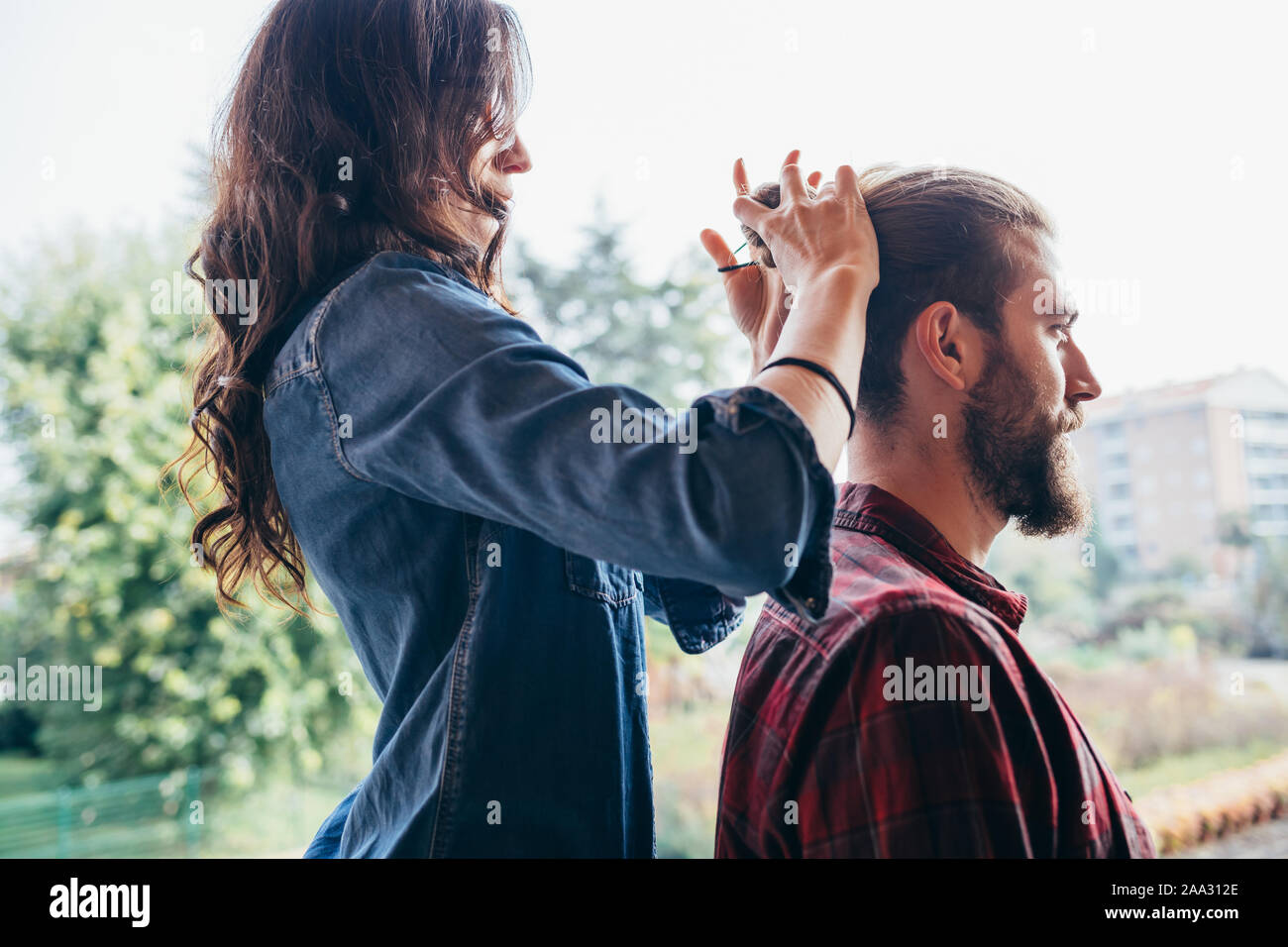 Mujer joven parado sobre una silla haciendo moño de cabello a su taller novio - ayuda, compañerismo, Care Concept Foto de stock