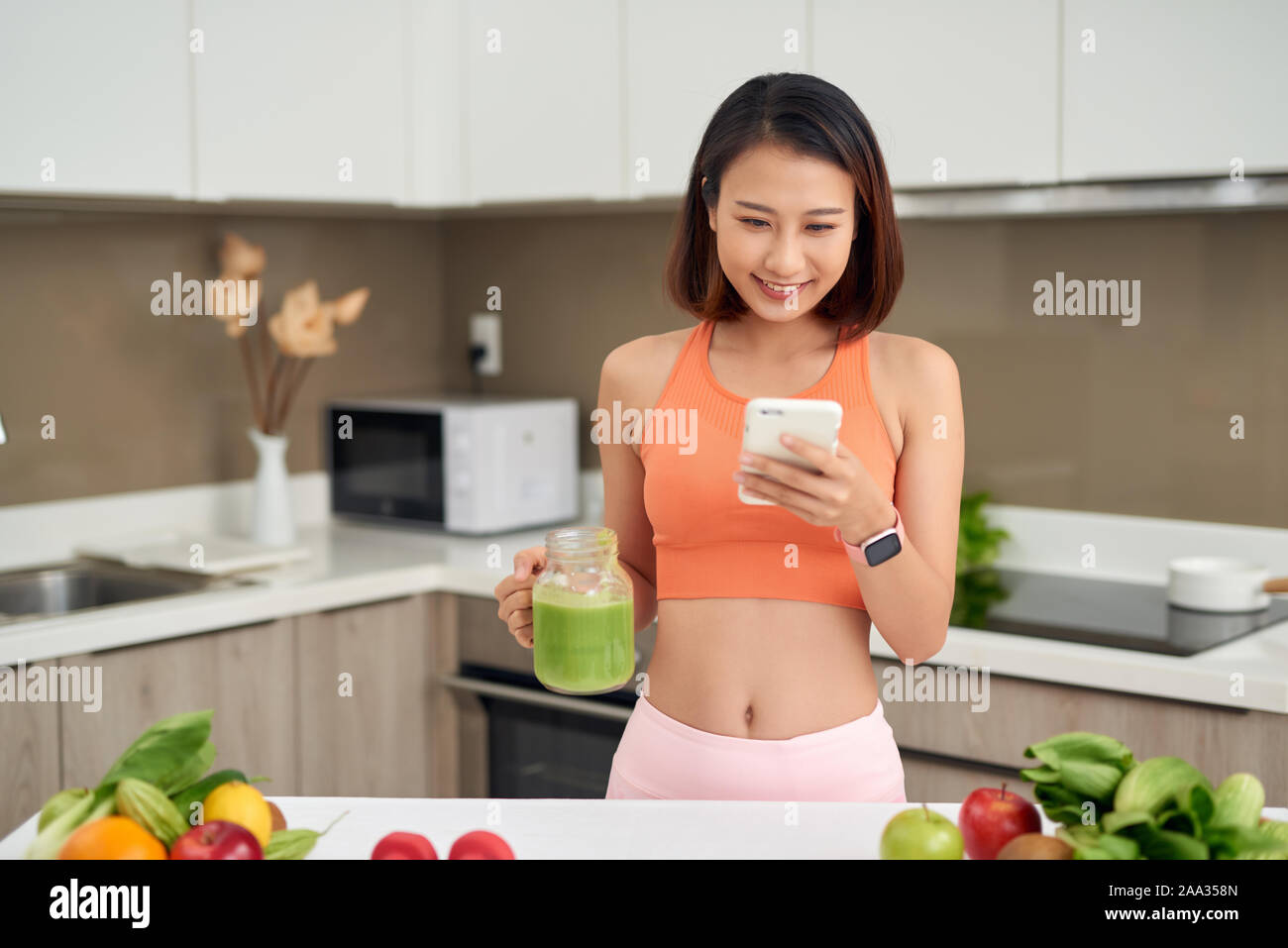 Mujer usando el teléfono mientras destintado smothie verde y de pie en la cocina Foto de stock