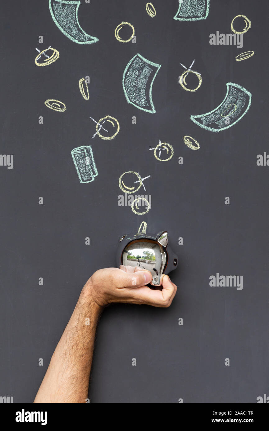 Concepto de ahorrar dinero con una hucha de plata celebrada en una mano delante de una pizarra con monedas y billetes dibujados a mano Foto de stock