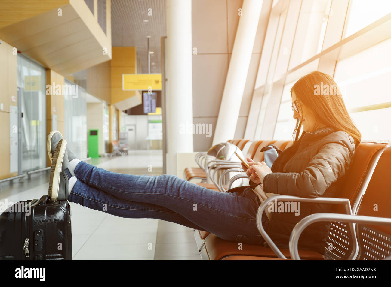 Mujer joven morenita está esperando en el aeropuerto para su vuelo, algo  que buscan en el smartphone. Ella se sienta con las piernas estiradas y  coloca en la maleta, la luz del