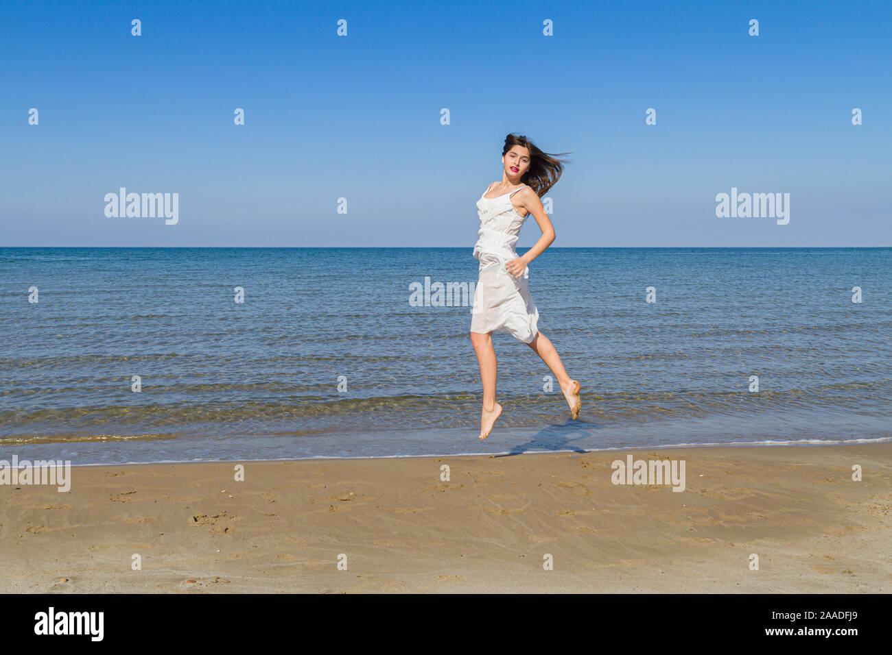 Joven hermosa niña despreocupada en endebles vestido blanco. La joven bella mujer alegre saltando sobre la playa en verano. Foto de stock