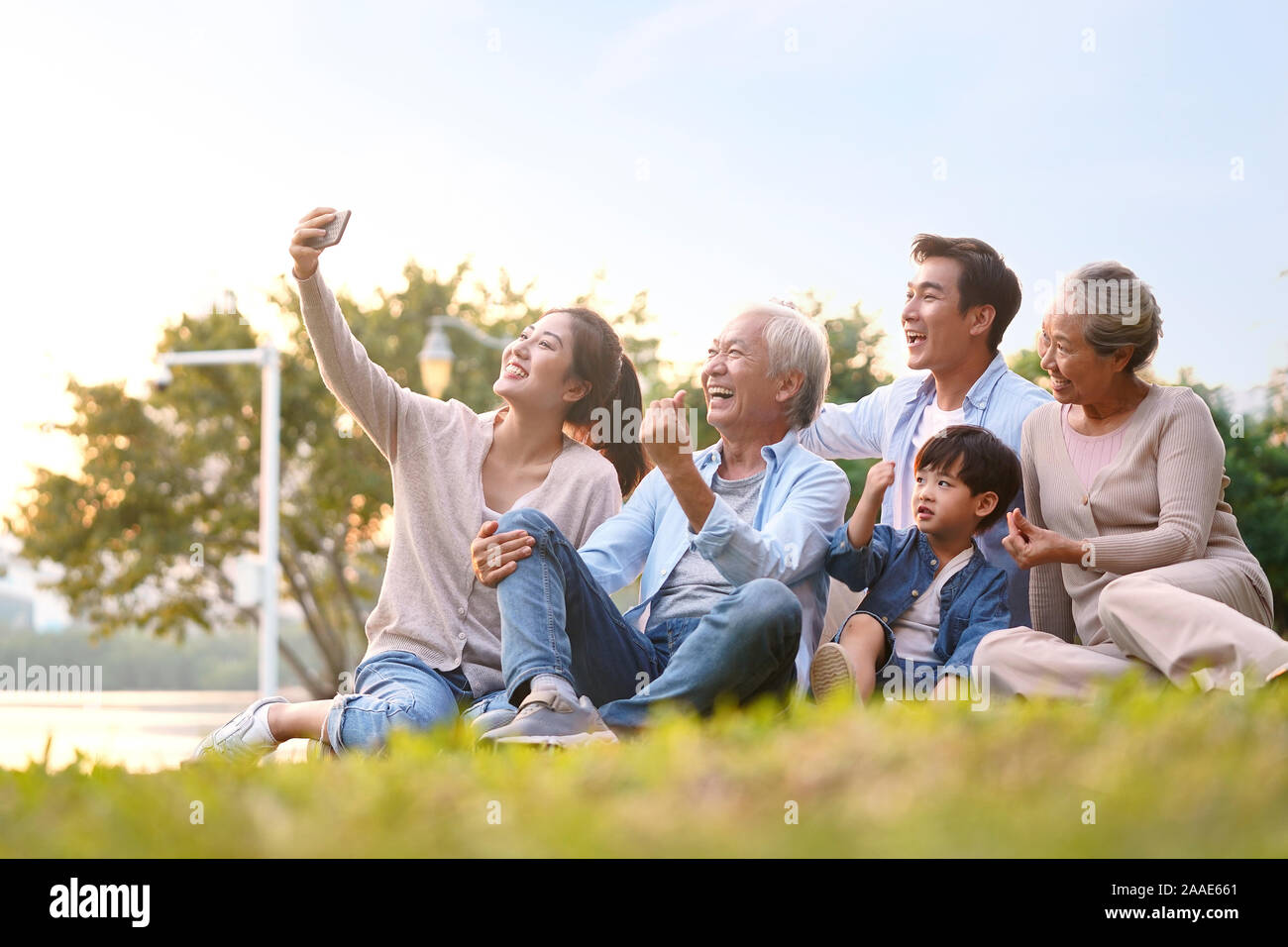 Tres generaciones de la familia de Asia felices sentados en el pasto tomando un selfie mediante teléfono móvil al aire libre en el parque Foto de stock
