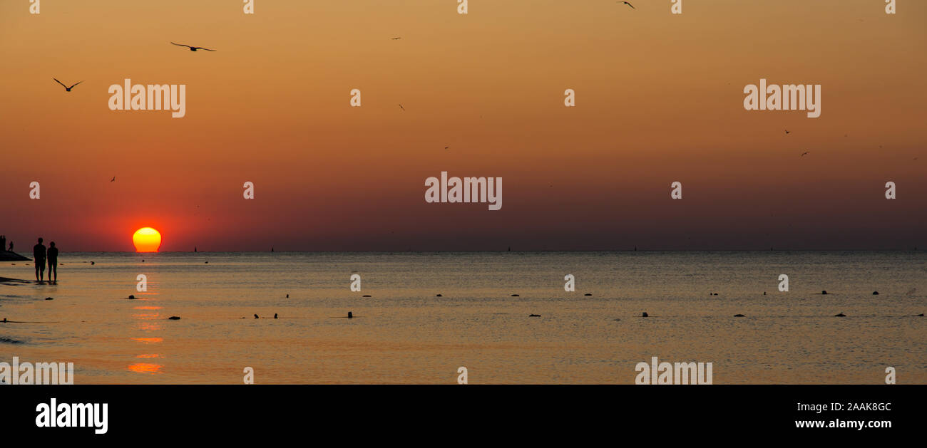 Romántica escena de siluetas de una pareja que ve el sol por la orilla del mar Foto de stock
