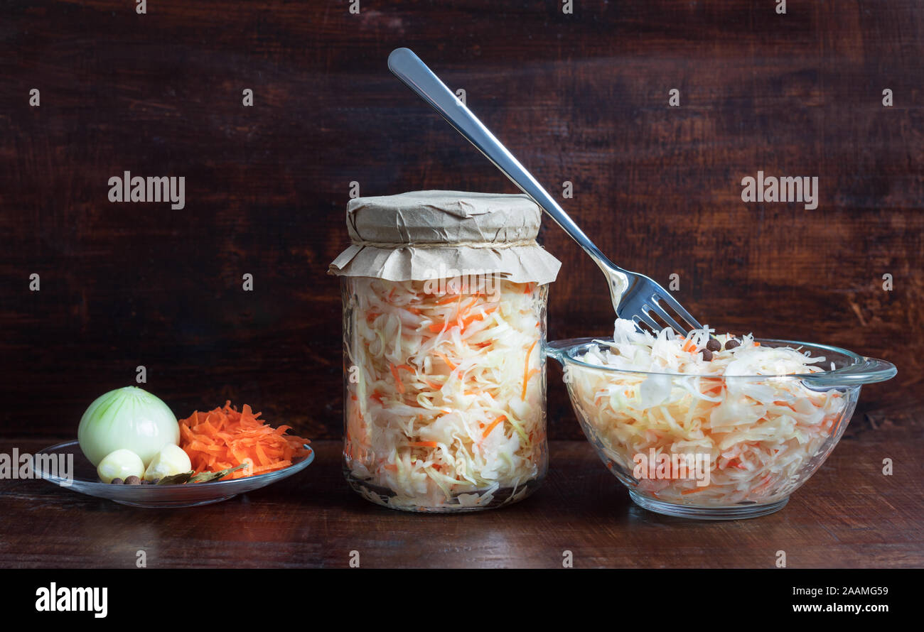 Chucrut casero con zanahorias en un frasco de vidrio y el tazón en un fondo de madera oscura. Alimentos fermentados. Verduras marinadas. Foto de stock