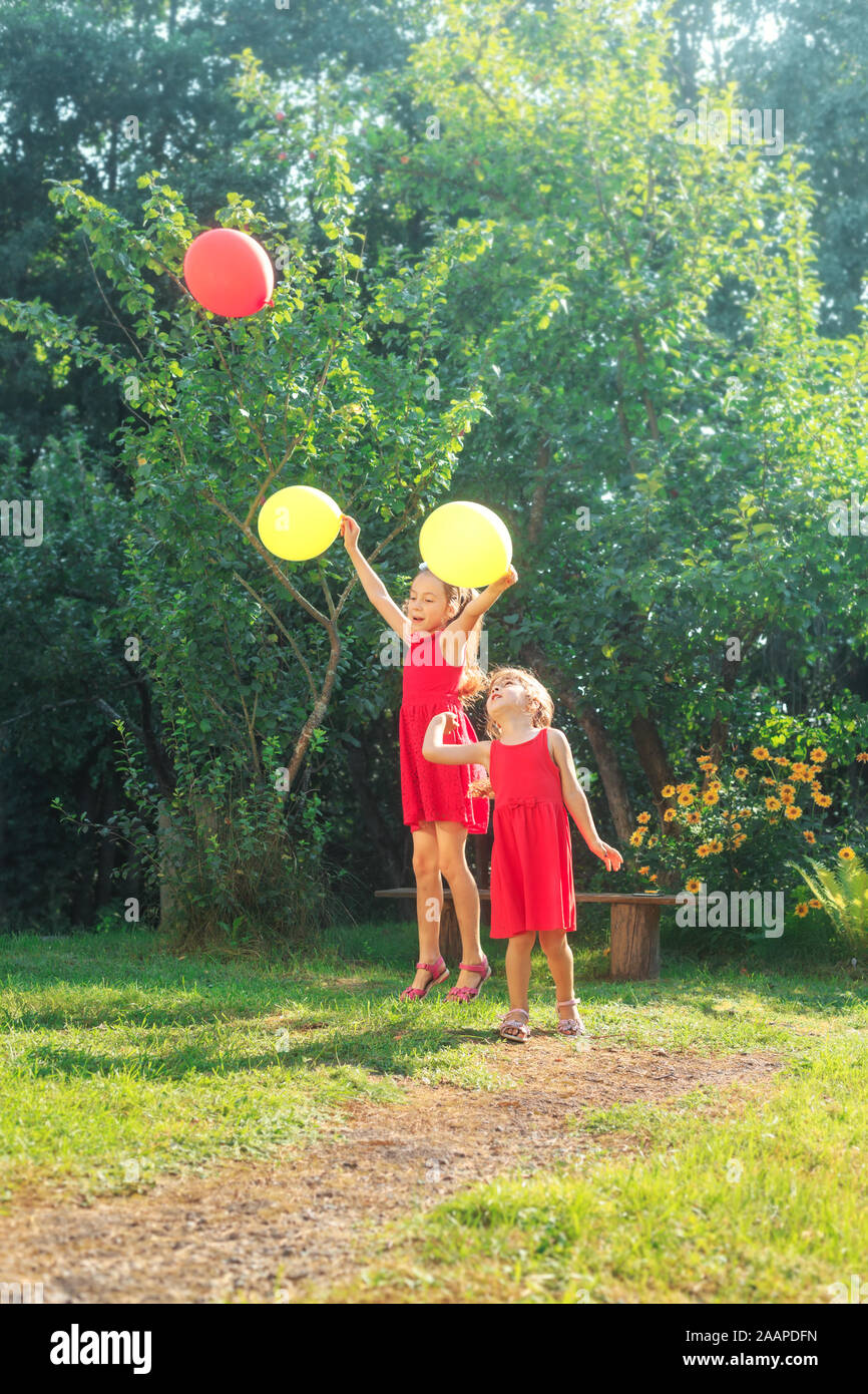 Dos hermanas cute little feliz saltando con coloridos globos de juguete al aire libre. Niños sonrientes divirtiéndose en el verde jardín de primavera en el día de verano. Foto de stock