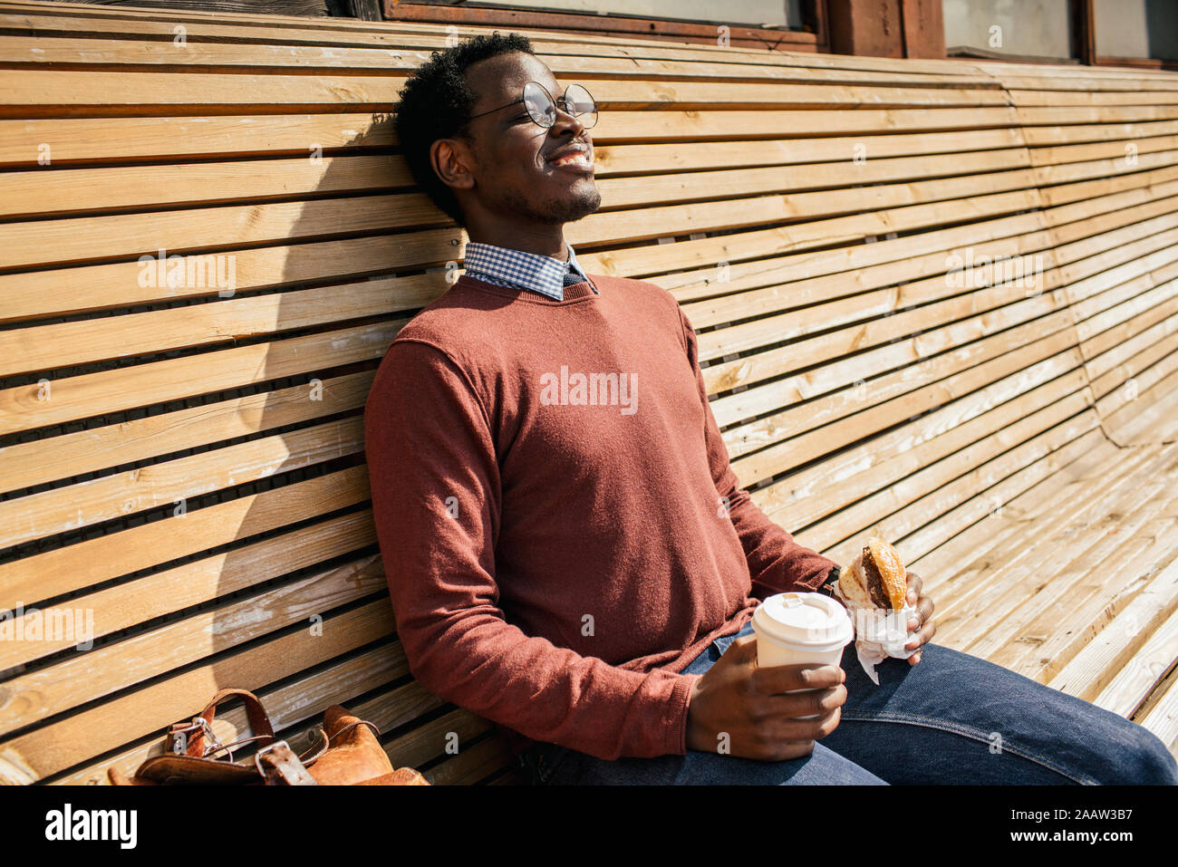 Hombre joven sentado en el banco de madera, comer hamburguesas, beber café Foto de stock