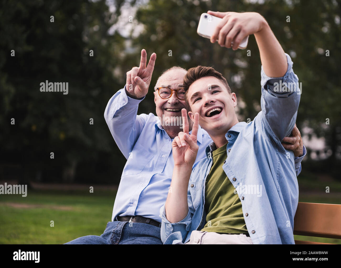 Hombre Senior y nieto sentados juntos en un banco del parque tomando selfie con el smartphone Foto de stock