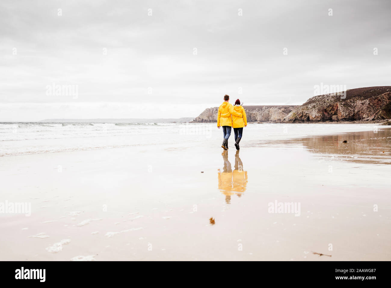 Mujer joven vestían chaquetas de lluvia amarilla y caminando por la playa, Bretagne, Francia Foto de stock