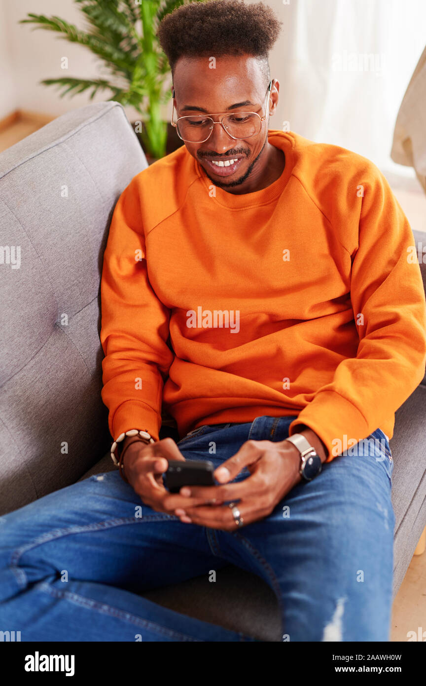 Sonriente joven hombre sentado en un sofá y utilizando el smartphone Foto de stock