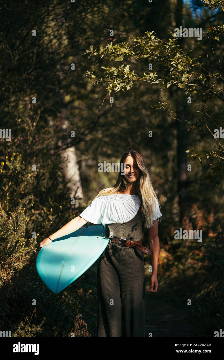 Joven Mujer rubia con un surfboard caminando en el bosque Foto de stock