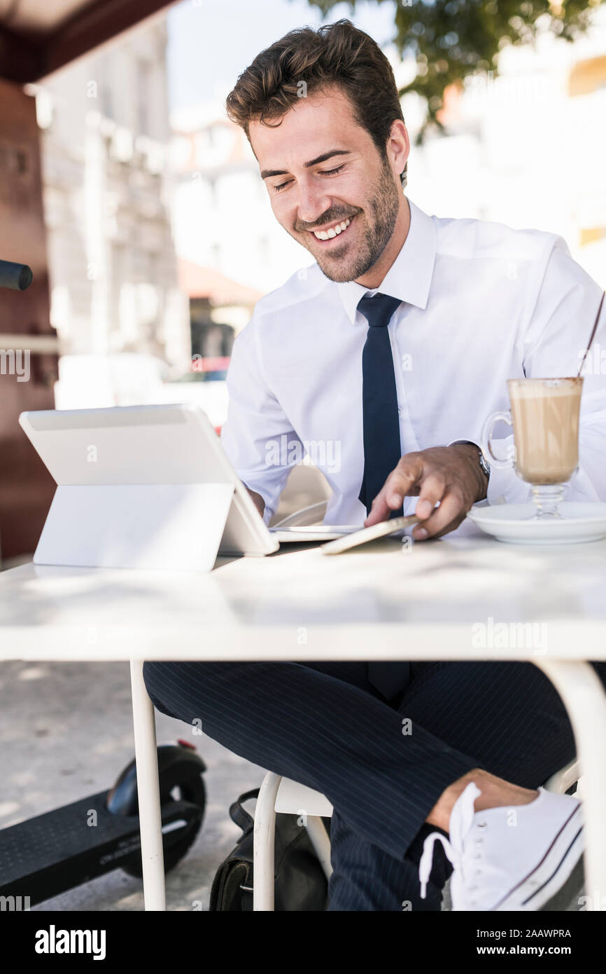 Sonriente joven empresario utilizando tablet y teléfono móvil en una cafetería de la ciudad, Lisboa, Portugal Foto de stock