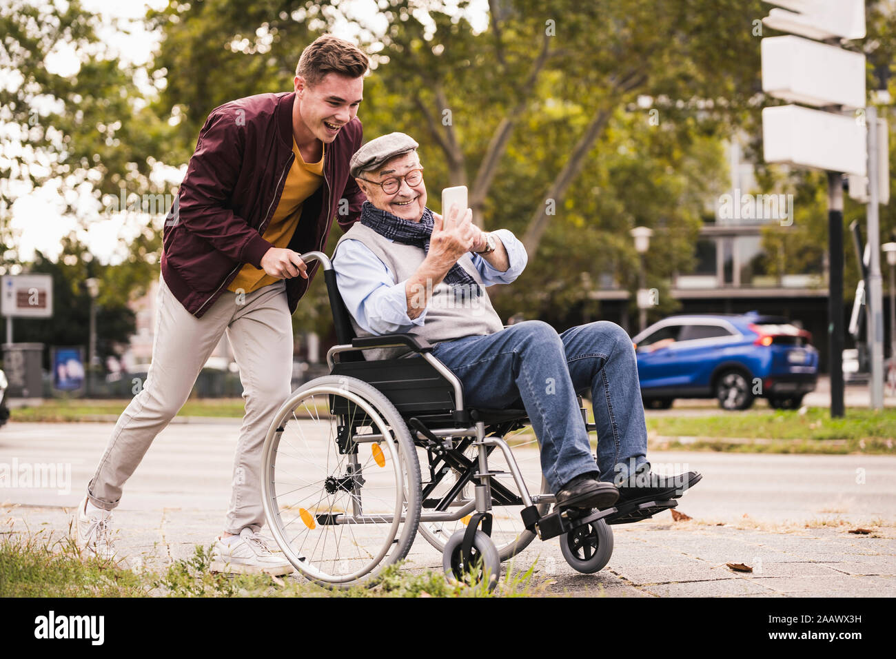 Sonriente joven empujando feliz hombre senior con el smartphone en silla de ruedas Foto de stock