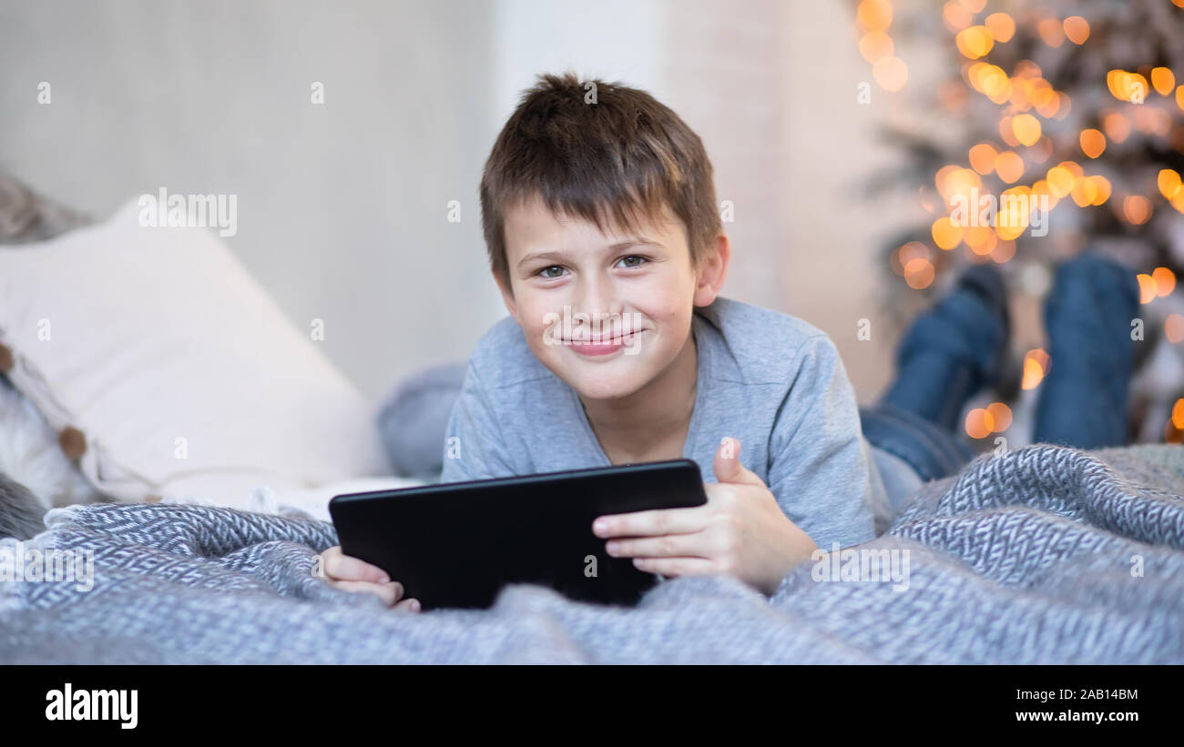 Feliz guapo muchacho con una tableta en manos. niño juega juegos de  ordenador en tableta. boy yace en la cama enfrente de árbol de Navidad antes  de Navidad. Frida negro Fotografía de