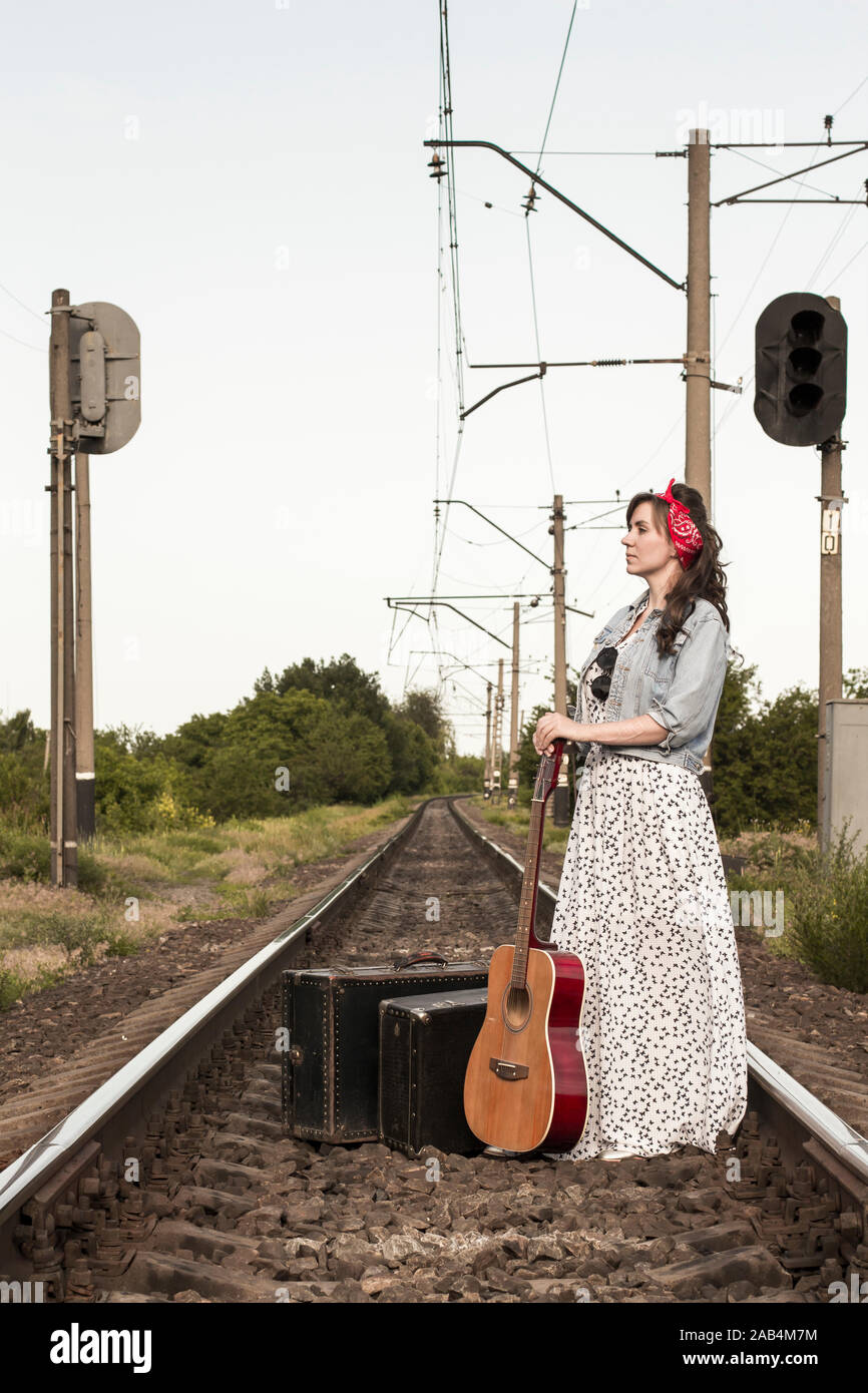 Hermosa chica con una guitarra. Pretty Woman con viejas maletas en las vías  del tren. Un adolescente en una chaqueta de denim, un largo vestido rojo y  un vendaje. Retro Fotografía de