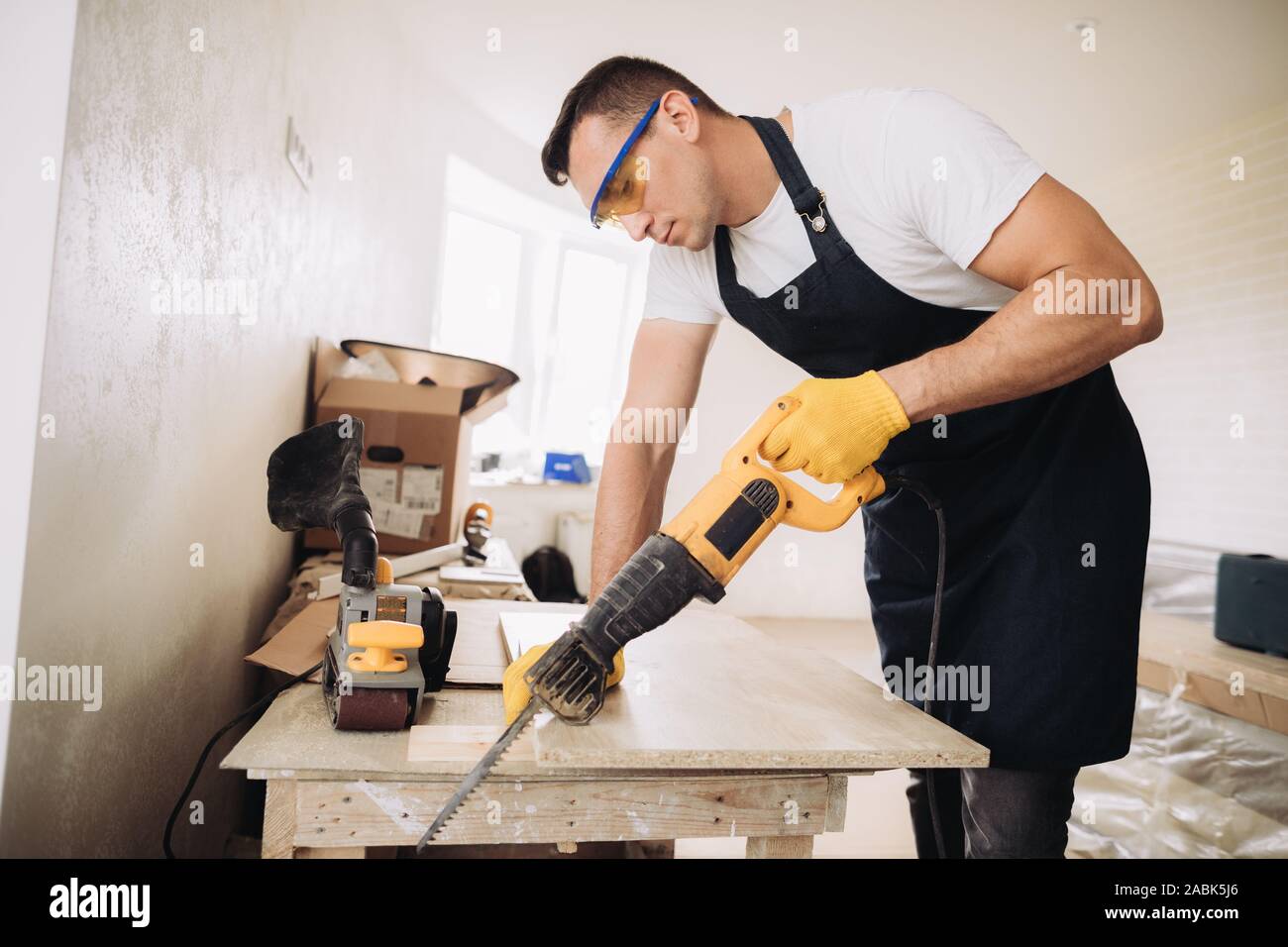 Hombre de mantenimiento está utilizando una sierra eléctrica para arreglar  los muebles en una habitación en construcción. Él está usando gafas,  guantes amarillos y un delantal negro Fotografía de stock - Alamy