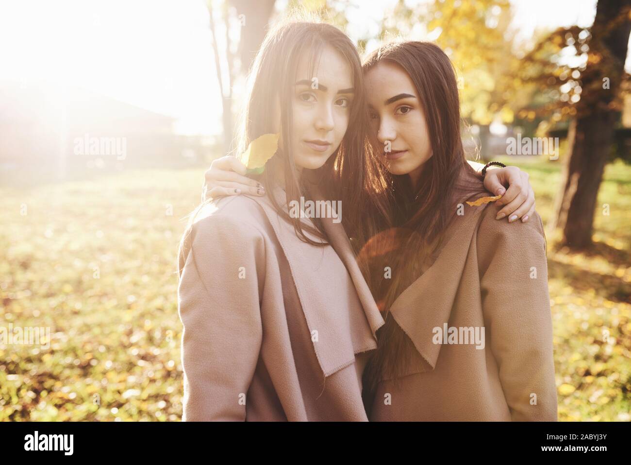 Dos niñas joven morenita muy cerca de pie se dirigió a cada uno de otros, tomados de la mano con las hojas sobre sus hombros, vistiendo chaqueta casual, tocando los jefes Foto de stock