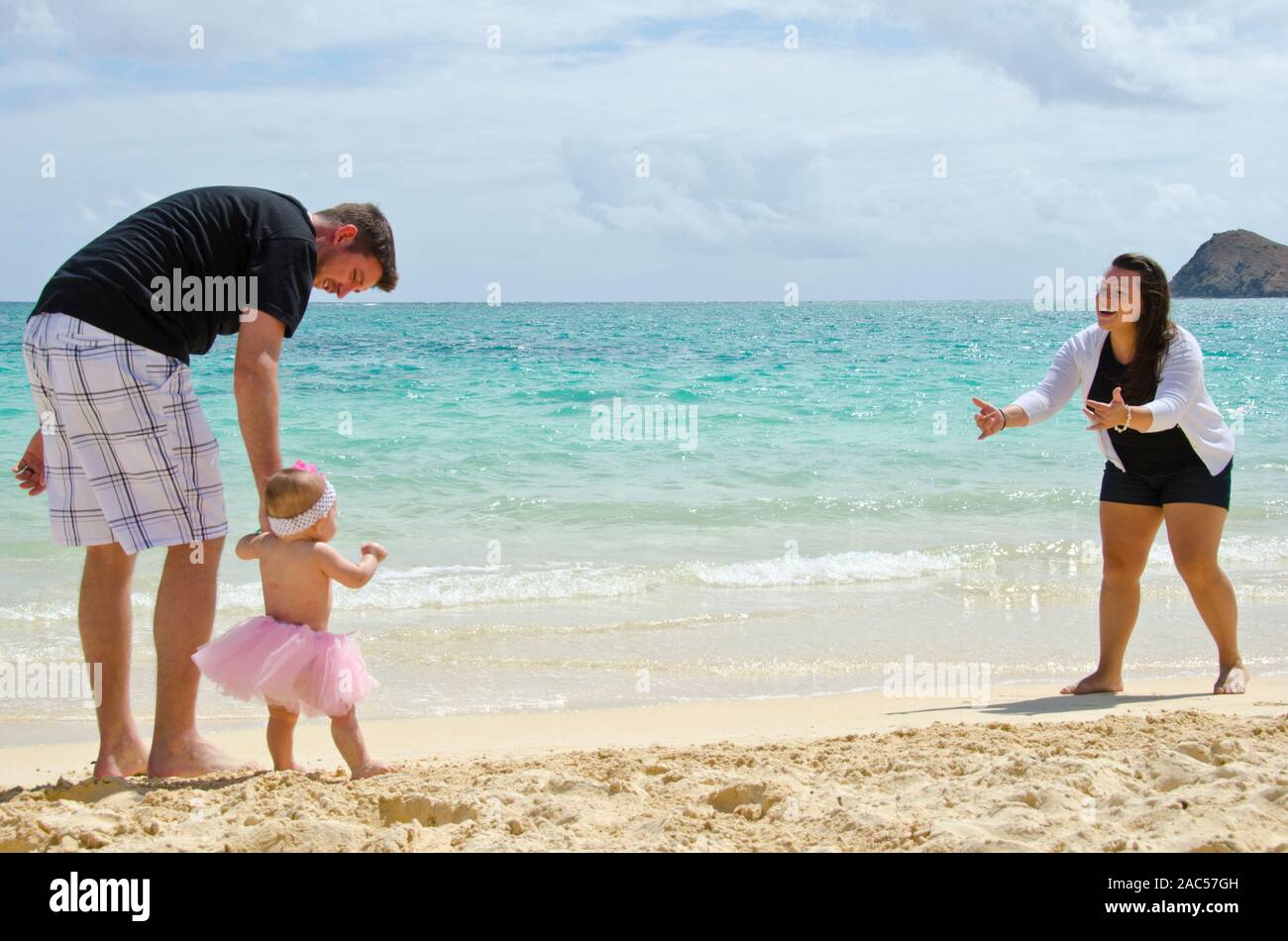 Una madre y un padre, alentando su joven hija a caminar a la orilla de la playa Lanikai, O'ahu. Foto de stock