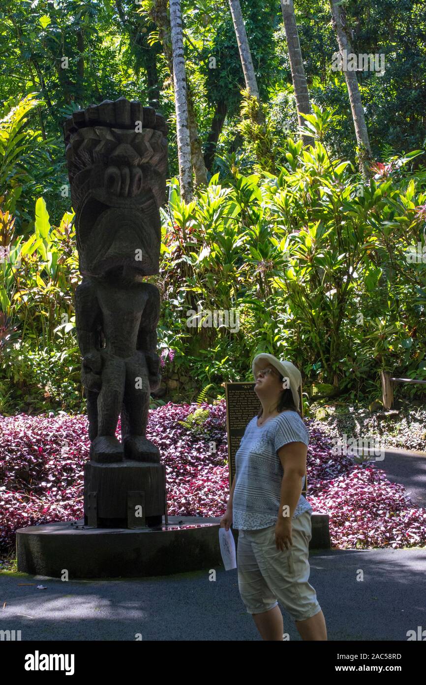 Una mujer mira hasta cerca de un tiki tallada en madera del dios hawaiano 'Ku' en el jardín botánico tropical de Hawaii, Papa'ikou, Isla Grande de Hawaiʻi. Foto de stock