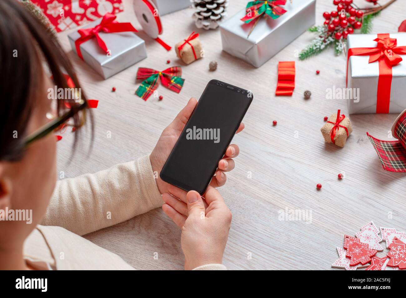 Chica sujetando un teléfono con pantalla en blanco para el boceto, rodeado por Navidad y Año Nuevo regalos y decoraciones. Foto de stock
