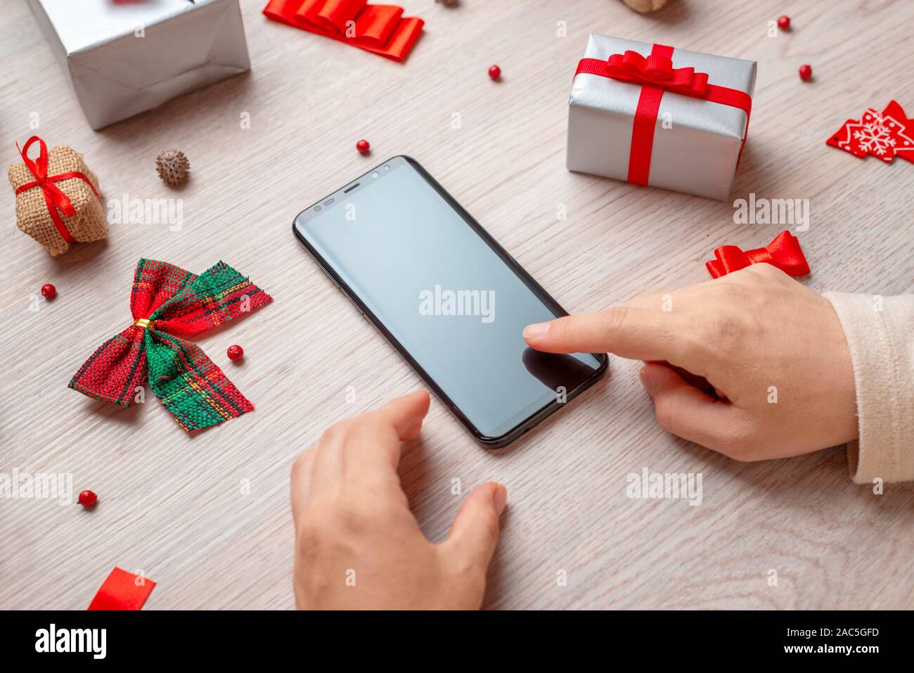 Maqueta de teléfono rodeado de adornos navideños y regalos sobre la mesa de madera. Chica en blanco pantalla táctil del teléfono. Foto de stock
