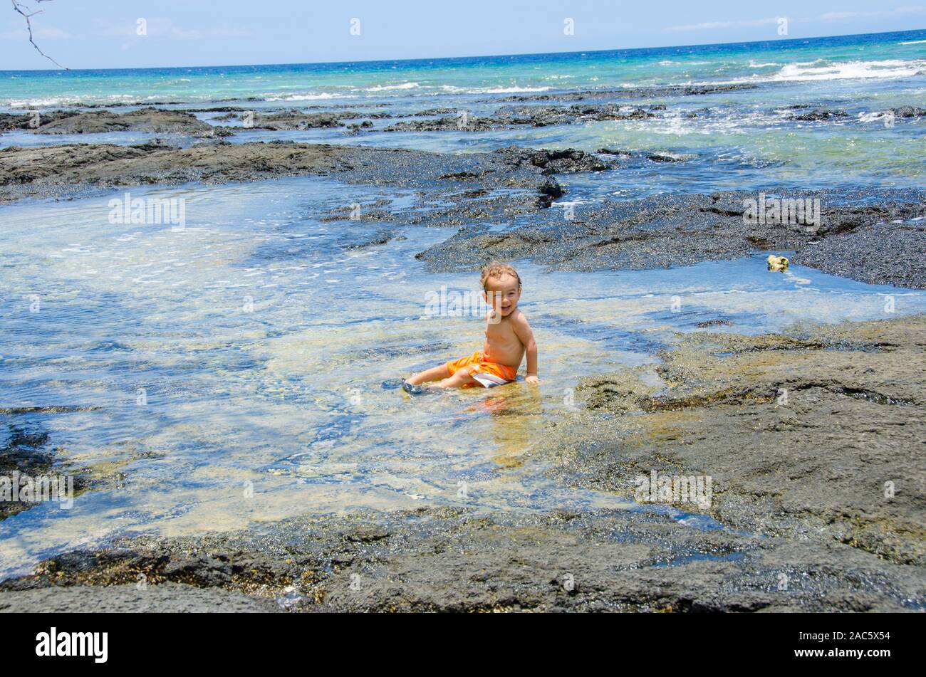 Un joven chico local se ríe como él se sienta en una piscina de marea en una playa en Puako, South Kohala, Hawai'i Isla. Foto de stock
