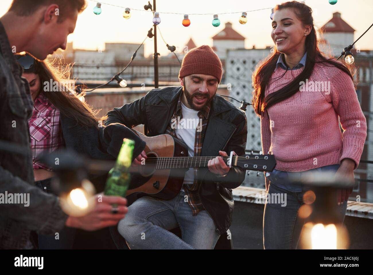 Jugar en el instrumento musical es siempre bueno tener habilidades. El guitarrista se sienta y cantando para sus amigos en la azotea con luz coloreada decorativos Foto de stock