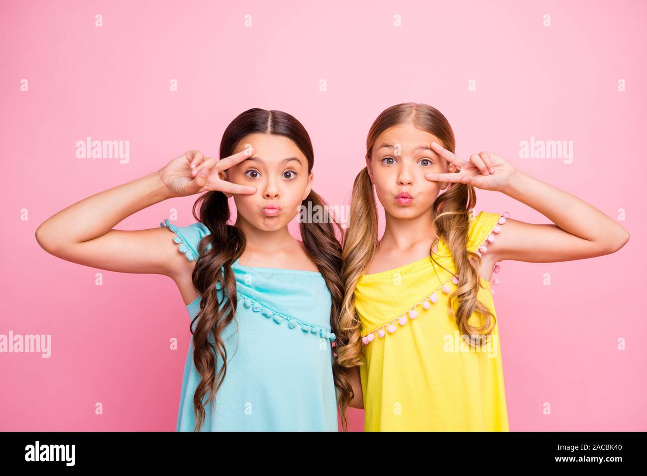Foto de dos pequeños modelos hermosas señoras amistad mostrando v-símbolos  de señal cerca de los ojos envía aire besos wear azul amarillo vestidos  color rosa aislado Fotografía de stock - Alamy