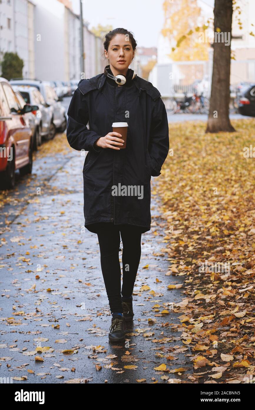 Mujer joven caminando por las calles de la ciudad solo en un frío húmedo  día de otoño vistiendo parka untar con auriculares alrededor de su cuello y  la taza de café para