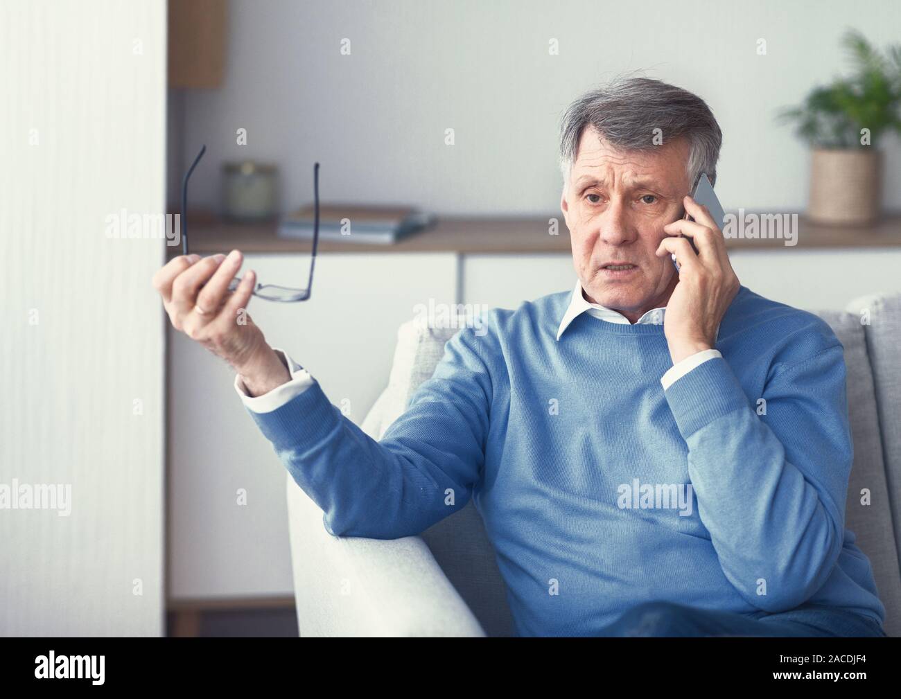 Hombre Senior tener conversación telefónica desagradable sentado en sofá para interiores Foto de stock