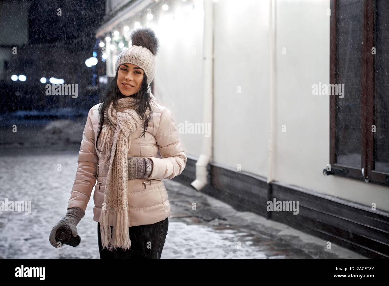 Hermosa niña positiva, situándose en calle con nieve. Señora vistiendo en  elegante y ropa de abrigo. Mujer joven en chaqueta, gorro de lana, bufanda  y guantes Fotografía de stock - Alamy