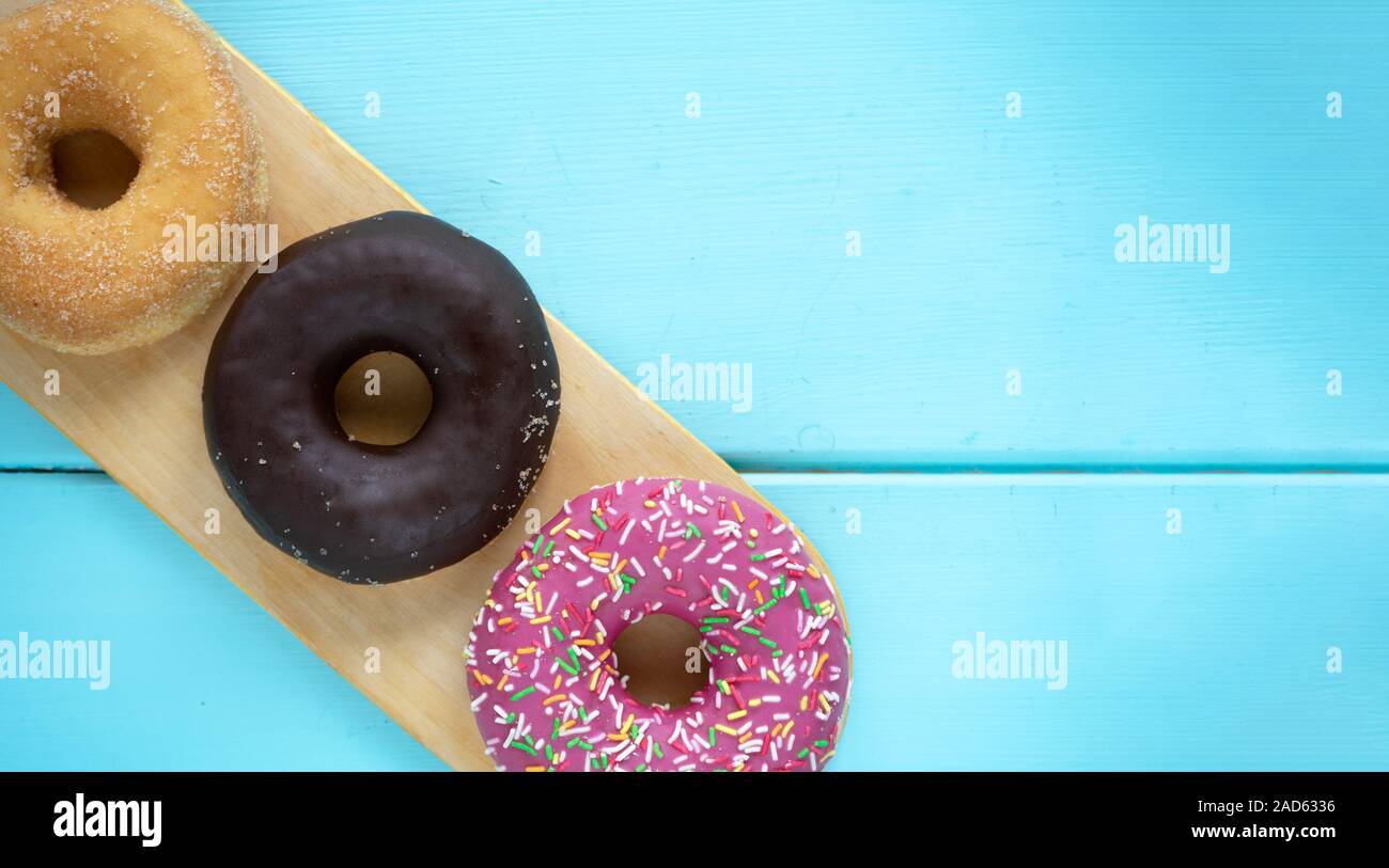 Tres diferentes donuts, chocolate donut, Rosa guinda y sin guinda sobre una tabla de cortar de madera de madera sobre un fondo azul. Foto de stock