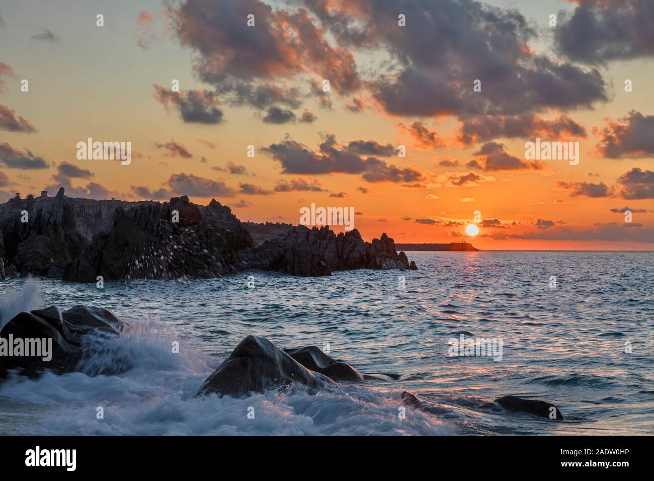 Atardecer con nubes y sol justo por encima del horizonte en Tropea desde playa con olas rompiendo sobre las rocas Foto de stock